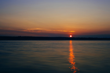 Naklejka premium sunset over river with sun and silhouette of the shore on warm summer evening