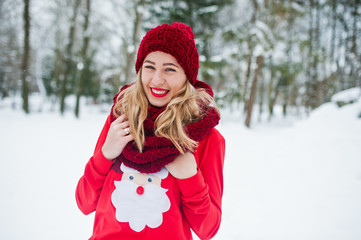 Blonde girl in red scarf, hat and santas sweater posing at park on winter day.