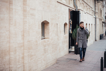 Fototapeta na wymiar Photo of elegant male in coat with bag in hand walking down empty street, and talking on smartphone