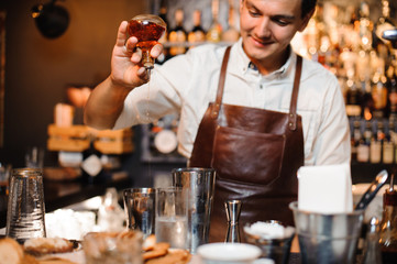 Young smiling barman in a brown leather apron making cocktail