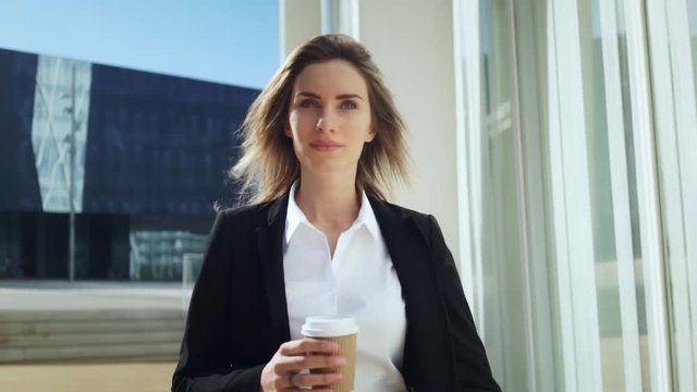 Portrait of young confident female ceo walking in sunny city, professional female lawyer wearing black suit drinking coffee at break, slow motion