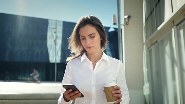 Beautiful young businesswoman wearing white shirt and using modern smart phone while walking at break in the city, professional female employer typing text message on cellphone outside, slow motion
