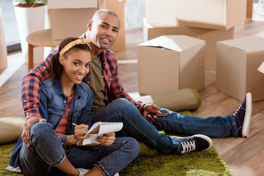 smiling african american couple with smartphone and notepad sitting in new apartment
