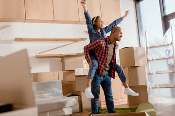 Fototapeta na wymiar excited african american couple piggybacking in new apartment with cardboard boxes