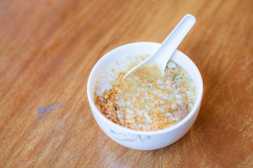 Boiled rice Asain Thailand style breakfast food in the morning, boiled rice wiht pork and deep fried garlic in the white bowl and white spoon put on the wood table