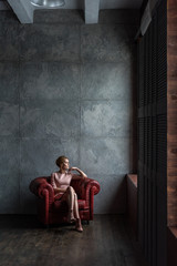 thoughtful young woman in pink dress sitting in armchair and looking at window