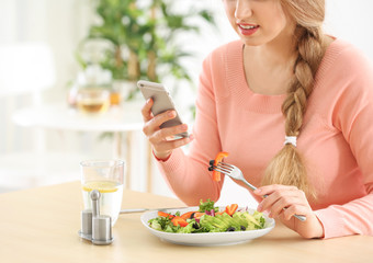 Young beautiful woman with phone eating fresh salad at table