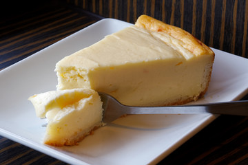 Plain baked cheesecake with cake on fork on white ceramic plate. Brown background.