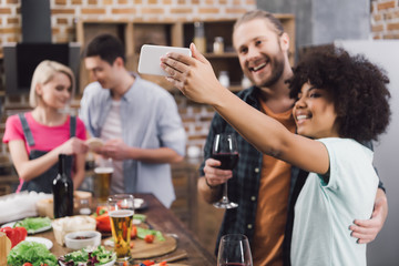 multiethnic friends taking selfie in kitchen with smartphone