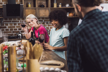 Obraz na płótnie Canvas multiethnic girls with wine looking at friends in kitchen