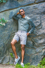 Young African American Man wearing long sleeve gray shirt, shorts, sneakers, sunglasses, standing against rocks at Central Park in New York, relaxing, thinking..