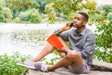Naklejka na ściany i meble Young African American Man relaxing at Central Park in New York. Young black man wearing gray long sleeves shirt, shorts, sneakers, sitting on rocks by lake, reading red book, talking on cell phone..