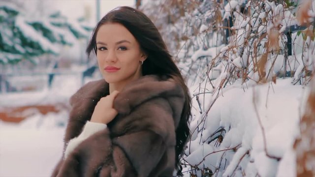 Close-up of Brunette fashionable sexy pretty rich woman or glamour model girl with beautiful long Brunette hair in waist coat of brown fur on background of tree posing for camera. slow motion