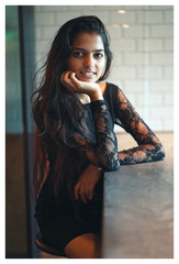 Portrait of young Indian Asian woman sitting at a cafe table, resting her face on her hand and smiling at the camera.  