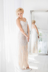 Beautiful young smiling white woman looking at camera posing at home interior. Beauty girl wearing gorgeous silver evening dress. Vertical color photography.