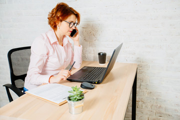 Red hair business woman in glasses working on computer and talking on phone in modern loft interior.