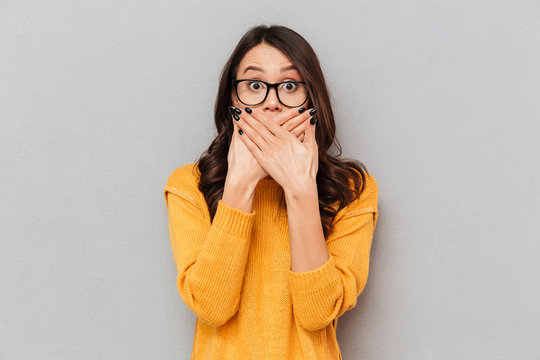 Shocked brunette woman in sweater and eyeglasses covering her mouth