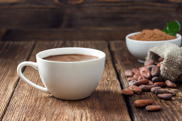 Cocoa drink in white mug, cocoa powder and cocoa beans on wooden table.
