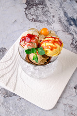 Top view of Dessert - Three balls of multi flavor ice cream with Scoops of Chocolate, Vanilla and Fruit Ice Cream with Fresh Mint in Glass Dish on a white plate on a gray background.