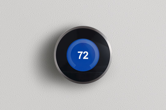 A simplistic photo of a round, modern, programmable digital thermostat in cooling mode, on a clean white wall.	
