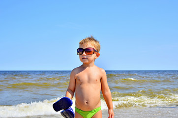 A boy in green swimming trunks with blue tapas in hands and brown glasses on the seashore