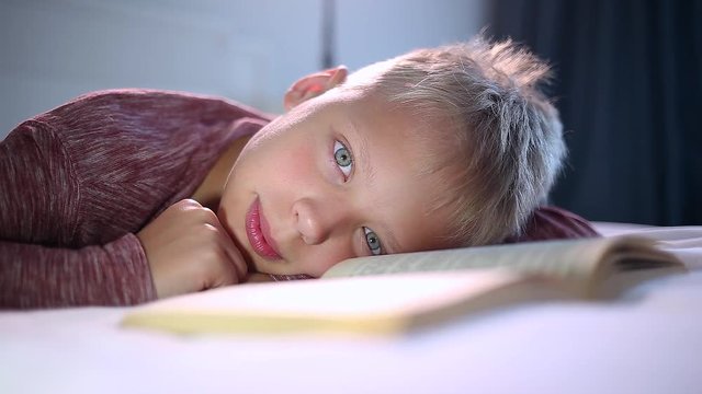 Closeup view of one little 10 years old kid reading paper book with interest laying in his bed at home.  Smiling cute white child looks at camera happily.