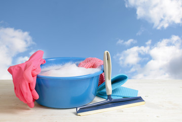 Spring cleaning, blue plastic bowl with soap foam, pink rubber gloves, rags and a window wiper on white wooden planks against a blue sky with clouds, copy space