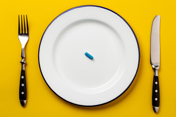 Blue pill on a white plate with fork and knife on a yellow background. Top view. Medical concept.