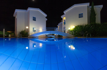 Бассейн с голубой водой и подсветкой в вечернее время, здания (корпуса) на заднем фоне