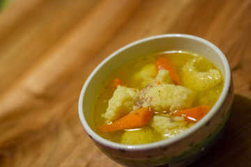 Hot chicken soup with dumplings and carrot in a small porcelain bowl on wooden table