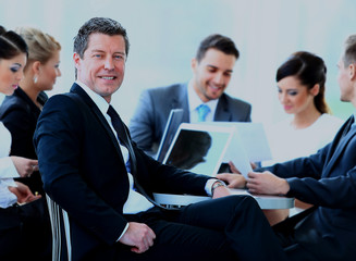 Portrait of mature business man smiling during meeting with colleagues in background.