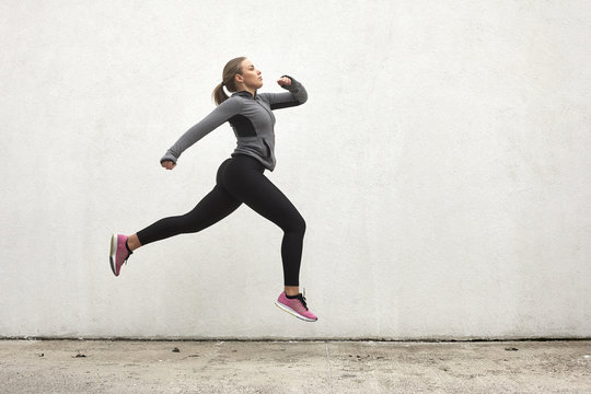One young woman, jumping in mid-air, outdoors, white wall behind, simple minimalistic, sport clothes.