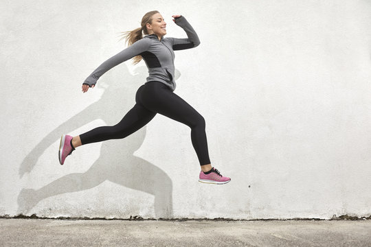 One young woman smiling happy, jumping in mid-air, outdoors, white wall behind, simple minimalistic, sport clothes.