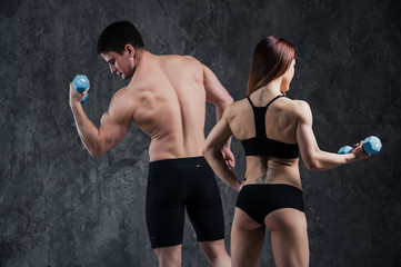 An attractive fitness couple of sporty male holds barbell and slim blond female holds dumbbells over grey background