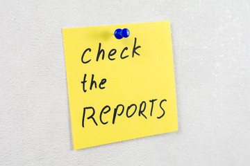 check the reports note reminder yellow sticker on a white wall pinned with blue pushpin, close up, selective focus