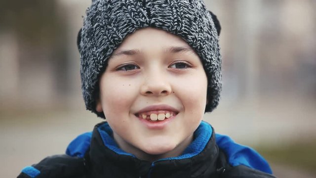 An emotional view of an excited boy standing on a city street on the Eve of Christmass. He wears a grey hat with black balls and smiles.