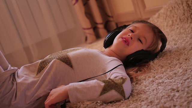 Cheerful young girl listening music in headphones lying on floor at home