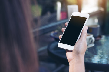 Mockup image of a woman's hand holding white mobile phone with blank black desktop screen and coffee cup in cafe