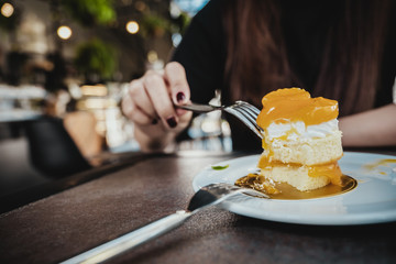 Closeup image of a woman cutting an orange cake with fork on white plate on wooden table in cafe