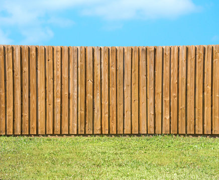 Generic wooden residential privacy fence with  a lush green grass yard in the foreground and a beautiful blue sky with fluffy clouds.