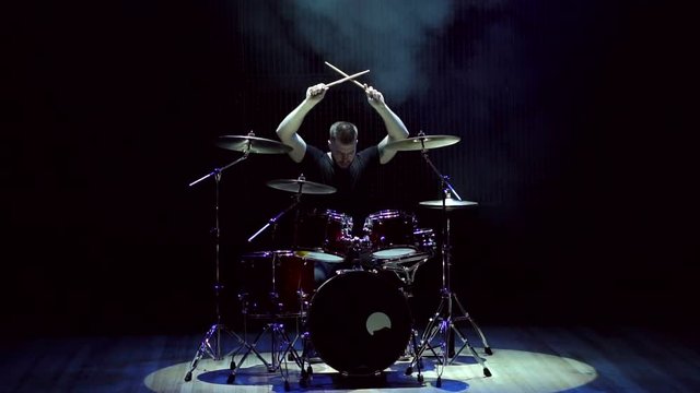 The drummer's man raised his hands with the drumsticks up and froze before the performance. The drummer sits behind the drum set on a black background before the concert.