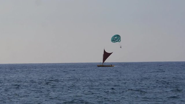 People parasailing pass a traditional Polynesian style sailboat