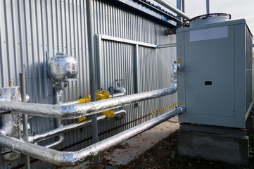 Insulated pipelines connecteed to the gray industrial cooling unit standing outdoor on the ground near to the modern fabrication building