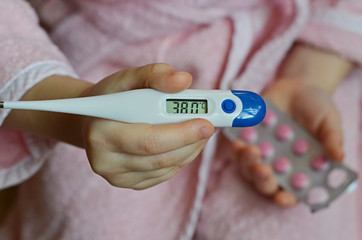 Little baby girl in pink bathrobe holding a digital thermometer with high temperature and pills in blister pack, close-up shot