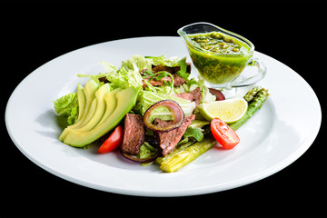 Delicious green meat salad with roasted juicy steak, avocado and green sauce in a white plate isolated on black background