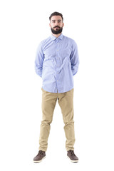 Fototapeta premium Front view of young smart casual bearded man standing with hands on back looking at camera. Full body length portrait isolated on white background. 