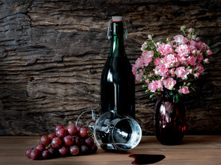 Still life visual art of grapes, a falled glass of wine, a bottle of wine and pink roses in a red vase on wooden slab with wooden back drop