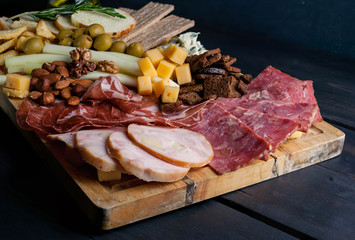 Assorted from meat and cheese on a chopping board. Smoked sausage, meat roll from a turkey with cheese, jamon, olives, nuts and various grades of cheese. Low key. Dark wooden background