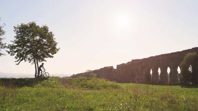 Cyclist riding bike uphill on dirt road in front of majestic ancient roman aqueduct. Young attractive athletic man with blue t-shirt and shorts sportswear in parco degli acquedotti in rome