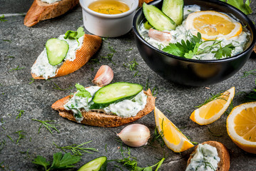 Traditional Caucasian and Greek food. Sauce tzatziki with ingredients - cucumber, lemon, parsley, dill, garlic. On a dark stone table. With sandwiches and baguette. Copy space